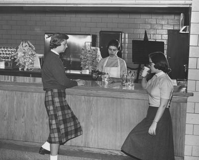 Martha Griffin (center) is pouring soda into a cup as Mary Adams (left) and Ann Fitzgerald (right) watch and wait at the concession in Keeneland Hall. Keeneland Hall was named after the Keeneland Foundation which donated $200,000 and on October 17, 1955, it was dedicated to the foundation. Received March 16, 1957 from Public Relations