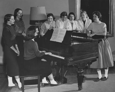 Seven unidentified women are standing around a piano and two unidentified women are sitting and playing the piano in Patterson Hall, UK's first woman dorm. Patterson Hall was completed in 1904 and was named after James K. Patterson. Received March 16, 1957 from Public Relations