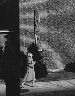 An unidentified woman is walking past the side of the Fine Arts Building, where there is a wooden sculpture on the building