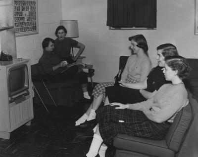 Three unidentified women of the Kappa Alpha Theta sorority are watching television and two unidentified women are talking, all are in the basement of the Kappa Alpha Theta House