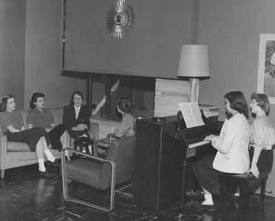 Four unidentified women of the Chi Omega sorority are talking while two unidentified women are playing the piano in the Chi Omega House