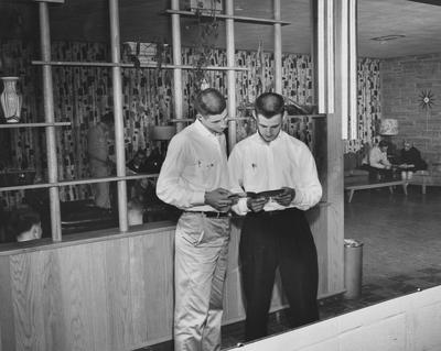 Two unidentified men are looking at a book with unidentified men conversing in the background, in the den of the Kappa Sigma house. Received March 18, 1957 from Public Relations