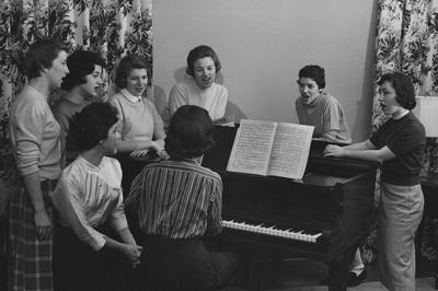 Carolyn Childers of Pikeville, Kentucky (second from right) and six unidentified women are singing around a piano while an unidentified woman is playing, in the Alpha Delta Pi house. Received March 19, 1957 from Public Relations