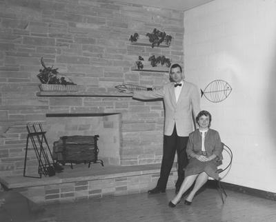 An unidentified woman is seated and an unidentified man is standing near a fire place in an unidentified fraternity house