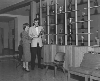 An unidentified woman is seated and an unidentified man are looking at a trophy in an unidentified fraternity house