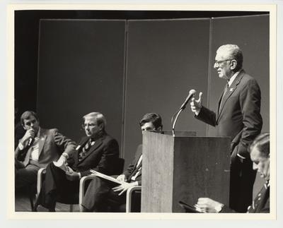 From the left:  State Senator Michael Maloney; University of Kentucky Alumnus Dudley Webb; State Senator Ernesto Scorsone; President Singletary (standing); and an unidentified man.  President Singletary is speaking at a podium to a group