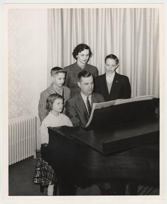 From the left:  Joe (standing); Betty (standing); Frank Dickey Jr. (standing); Ann (sitting); and President Dickey (seated).  The family is gathered around a piano.  Received by Public Relations on August 17, 1957