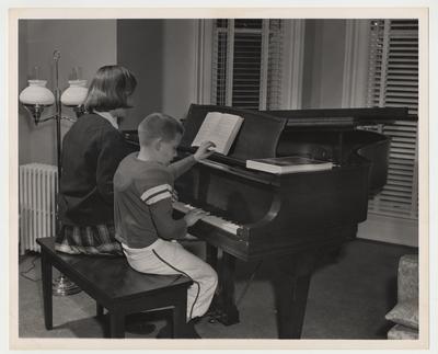 John Oswald Jr. in his football uniform is practicing piano at Maxwell Place.  The woman playing piano with him is either his sister Nancy or an unidentified piano teacher