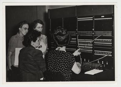 Four women are in front of a switchboard.  Standing from the left:  Elizabeth Holtzclaw (part time operator) and Betty Swope (chief operator.)  Sitting from the left:  Carol Reeves ( University operator) and Ms. James 