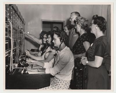 Several unidentified women are operating the new switchboard while other unidentified men and women watch