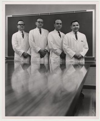 Four unidentified men in white coats are standing in a room