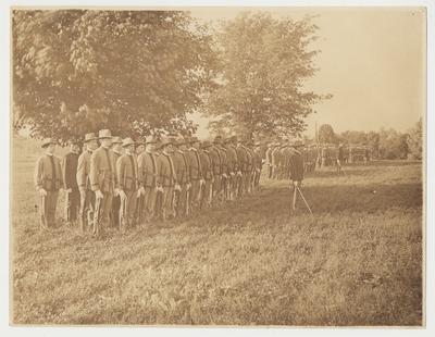 Spanish American War Era troops in formation wearing uniforms and carrying guns.  The commanding officers are in front with swords