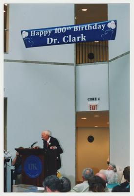 Loretta Brock (seated, right) and unidentified people are listening to Thomas Clark (standing, left) speak at Dr. Thomas D. Clark's 100th birthday celebration at Young Library