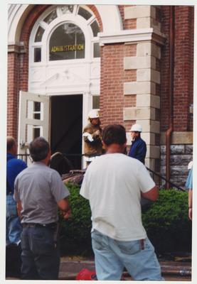 President Wethington (right, blue coveralls and a white hat) and three unidentified men are standing outside of the Administration / Main Building during the fire.  Wethington is talking to a fire fighter