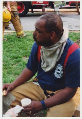 An unidentified, male, African American fire fighter is seated on the ground and eating after helping to extinguish the fire in the Administration / Main Building