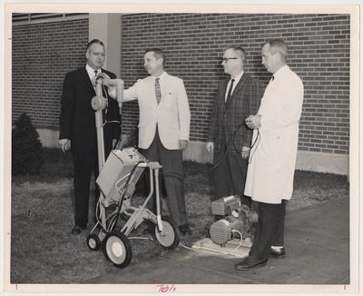 Four men are standing with a portable x-ray machine.  From the left:  Lawrence Hill, Chief X-Ray Technician, State Health Department; Dr. H. M. Vaniviere, Associate Professor of Community Medicine; Thomas Layton, former secretary of the Kentucky Hospital Commission; and Luther Hall, x-ray technician