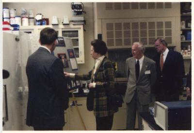 A small group of people faces an unidentified man who is standing in front of a display of photographs.  From the left:  an unidentified man; Peter Timoney, director of the Gluck Equine Research Center; the Princess Royal Anne of Great Britain; an unidentified man; and UK President Lee Todd
