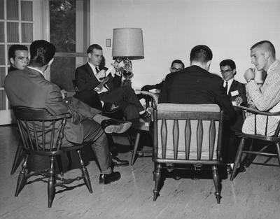 Seven men seated in a circle, and one is playing a ukulele