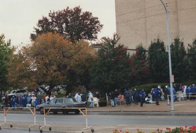 A color photo of unidentified people standing and sitting in line as well as two officers on horses at Memorial Coliseum for 
