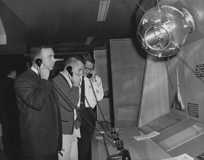 Three men listening on phones at a display at the 1960 Career Carnival. Received November 1, 1960 from Public Relations