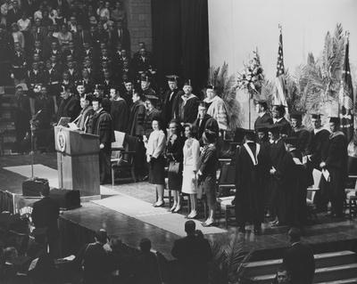 Centennial Convocation platform party. Featured speaker was President of the United States Lyndon B. Johnson (far left on platform head bowed). At podium is Kentucky Governor Edward T. Breathitt. To his right stands UK President John W. Oswald. Women standing (left to right) are: Lady Bird (Mrs. Lyndon B.) Johnson, Rosanel (Mrs. John W.) Oswald, Mrs. Edward T. Breathitt, and one unidentified woman