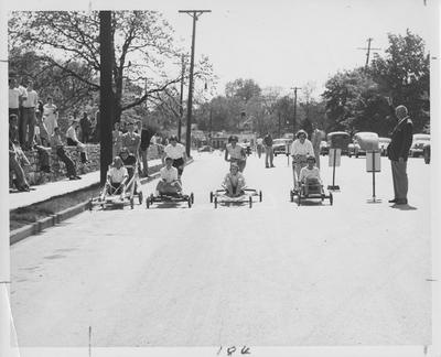Lambda Chi Alpha pushcart derby. Women seated in carts (from left to right) are Pat McDeavitt, unidentified, unidentified, Greta Boswell and man standing on far right is Mr. Bernie Shively