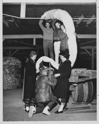 Homecoming Float being constructed in a barn or tobacco warehouse. Three men and three women working on the trailer. Woman in wool coats with raccoon collars