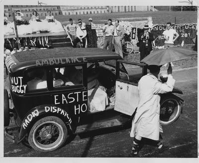 At Stoll Field, Homecoming Court practice pre-football game. Women students on court are on a platform with formals and bouquet of flowers. Man with trumpet and man with camera and other men watch an old 1920's car that is part of homecoming activities. Persons dressed to appear insane and car is the 