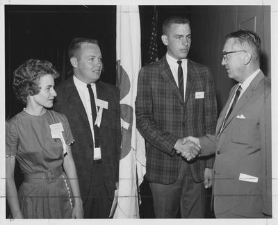 One unidentified male 4-H member shaking hands with an unidentified 4-H speaker. Unidentified members are standing near by