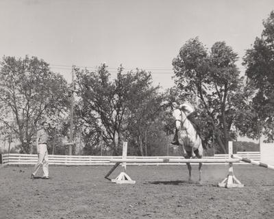 Barbara Harper on horse jumping an obstacle while Robert Garrigus watches. Received May 17, 1958 from Public Relations