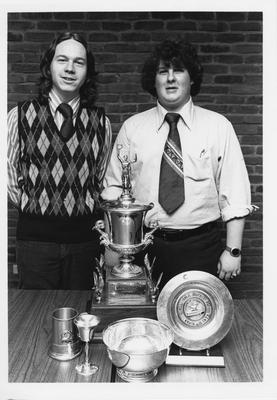 Two unidentified men standing with five trophies