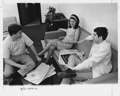 Three unidentified graduate students (and a cat) seated on couches and looking at newspapers and talking. This image appears second on page 204 in the 1969 Kentuckian