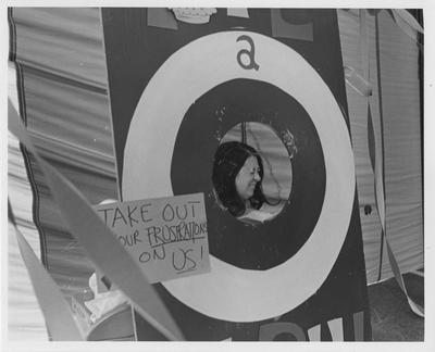 Girl seated at a pie-throwing booth; Sign says 