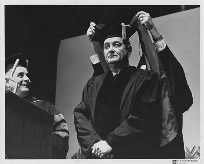 United States President Lyndon B. Johnson, speaker at Centennial Convocation (1965, February 22), receiving an honorary degree; Placing an academic hood on the President is Kentucky Governor Edward T. Breathitt; Looking on at left, at podium, is University of Kentucky President John W. Oswald; Photographer: R. R. Rodney Boyce