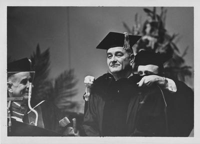 United States President Lyndon B. Johnson, speaker at Centennial Convocation (1965, February 22), receiving an honorary degree; Placing an academic hood on the President is Kentucky Governor Edward T. Breathitt; Looking on at left, at podium, is University of Kentucky President John W. Oswald; Lexington Herald - Leader staff photo