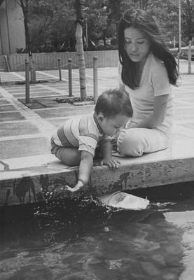 A woman watches as a child plays with a toy boat in the fountain