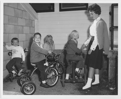 A woman stands with children on tricycles