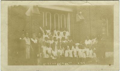 Group photo of unidentified African American children in uniform and adults standing on the steps of a large building