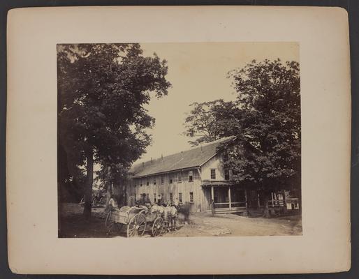Office of the quartermaster and commissary; large wooden building with porch and two floors, three men riding in wagon pulled by horses while other men stand in front of building, dirt road and trees in foreground, trees in background
