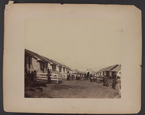 Refugee camp; Street scene; dirt road with small white wooden houses lining both sides far into the distance, crowd of African-American women and children posing in street around man on horse