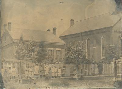 Little girls standing in front of a picket fence surrounding two buildings-two men standing at the gate