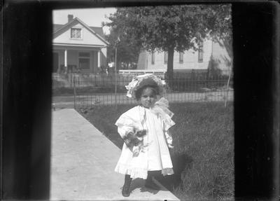a young child (appears to be the same child as in image #3) standing on the front walkway wearing a dress and bonnet