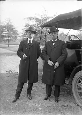 2 men wearing coats and hats, standing next to a car