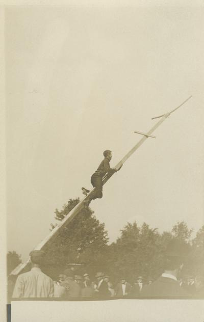 an unidentified man climbing up a pole, others watching him circa 1910