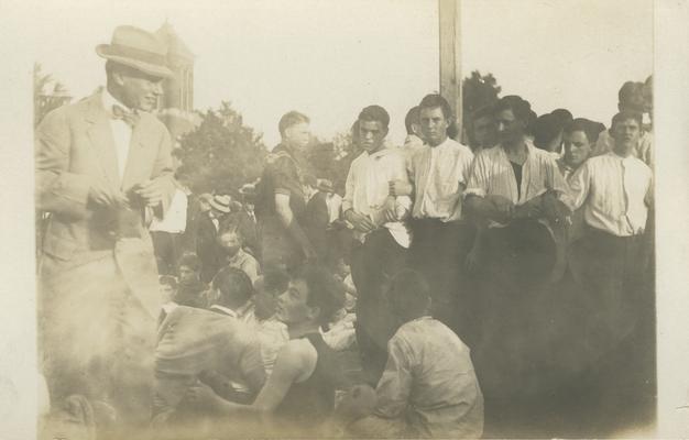 a group of unidentified men protecting the flag pole by linking arms, other unidentified men around their feet, 1 unidentified man wearing a suit and talking to group circa 1910