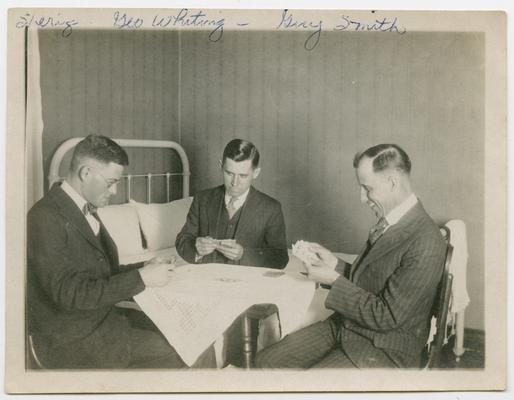 Sherring?, George Whiting, Gary Smith playing cards sitting at a square table covered with a cloth.  There is a bed in the background