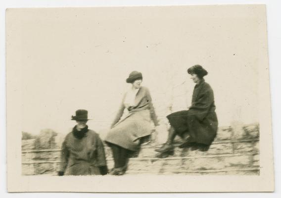 Two women wearing hats and coats sitting on a rock wall with a third woman also wearing a hat and coat standing next to them
