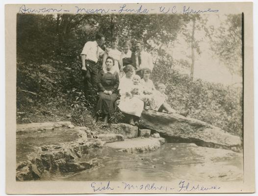 Dawson, Mason, Linda, W.C. Grimes, Eich, Mrs. Olney, Florence sitting with two unidentified children on a rock by a stream.  There are trees in the background
