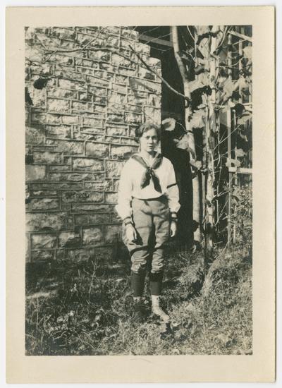 Ethel Anderson from New York state, wearing a sailor shirt, breeches, and boots, standing in front of a stone wall