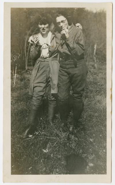 Two unidentified women smoking cigarettes.  They are wearing breeches, leggins, and boots with long-sleeved shirts